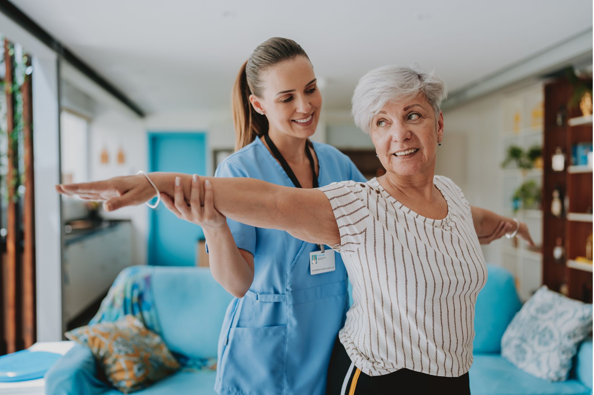 A healthcare professional helps an older woman extend her arms out to the sides while standing in a living room, both smiling during a guided movement exercise.