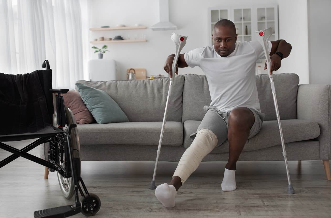 Man with a bandaged leg using crutches during home recovery and rehabilitation after a lower leg injury, with a wheelchair nearby.