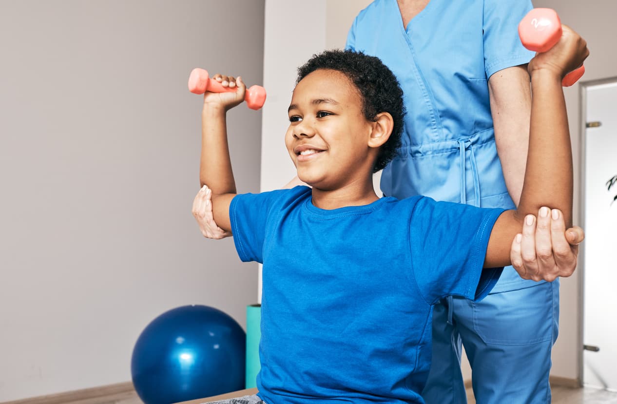 Young boy performing arm strengthening exercises with dumbbells while assisted by a pediatric physical therapist during a rehabilitation session.
