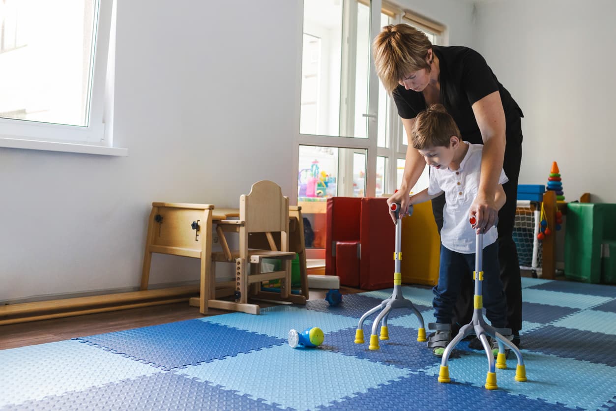 Pediatric physical therapist assisting a young boy using mobility walkers during a rehabilitation session in a therapy room.
